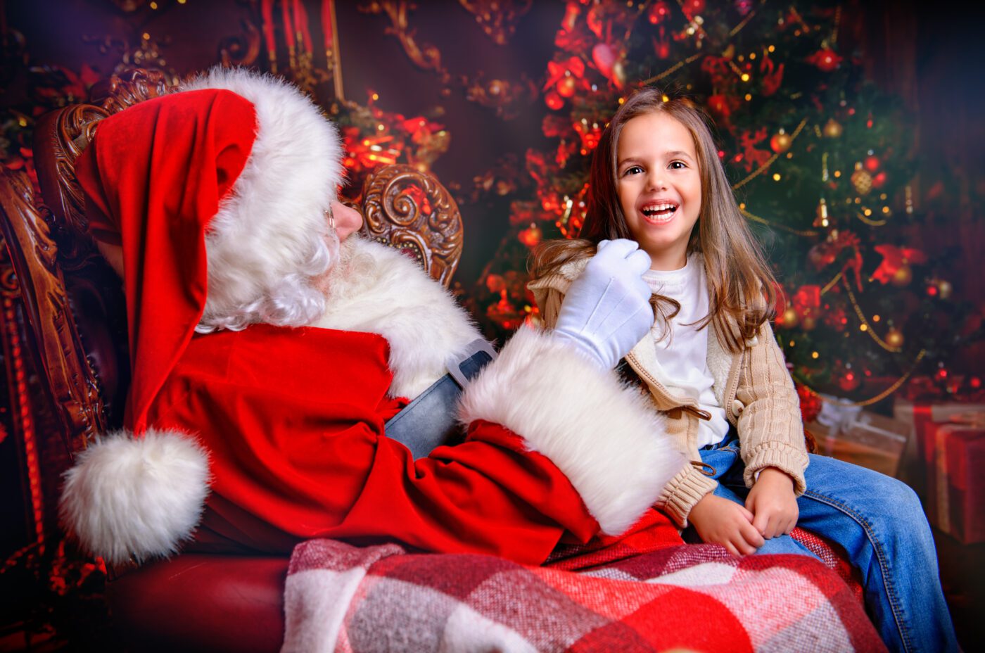 A young girl sits on Santa Claus’s lap, smiling and laughing in a festive room reminiscent of the Taste of London, decorated with a lit Christmas tree and red ornaments. Santa is dressed in his traditional red suit and white gloves.