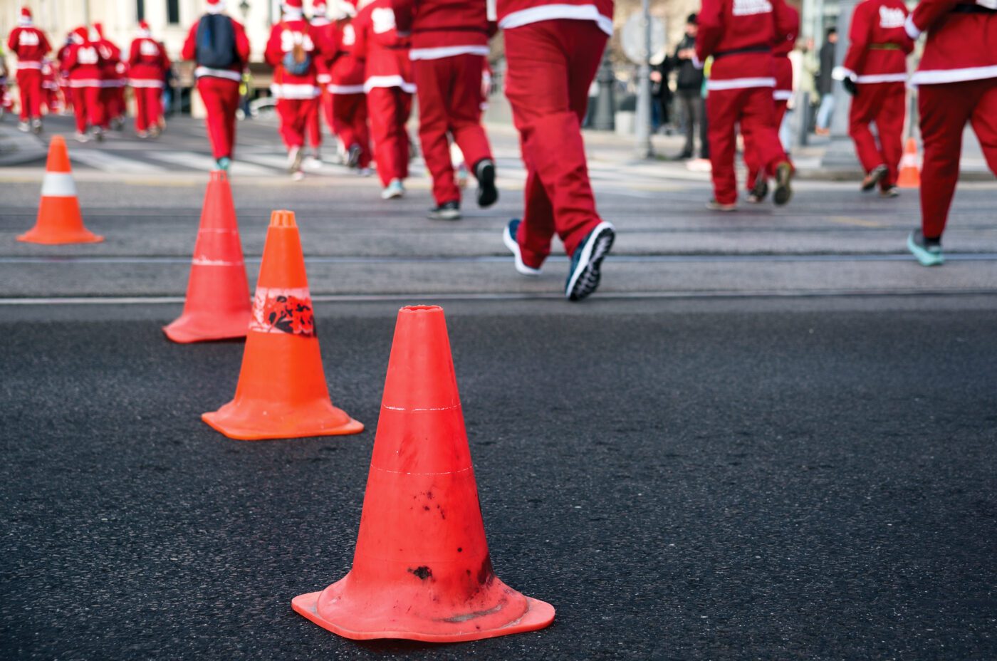 Several people dressed in red Santa Claus costumes are running on a city street during the biennial event, with orange traffic cones in the foreground marking the path. The photo is taken from a low angle.