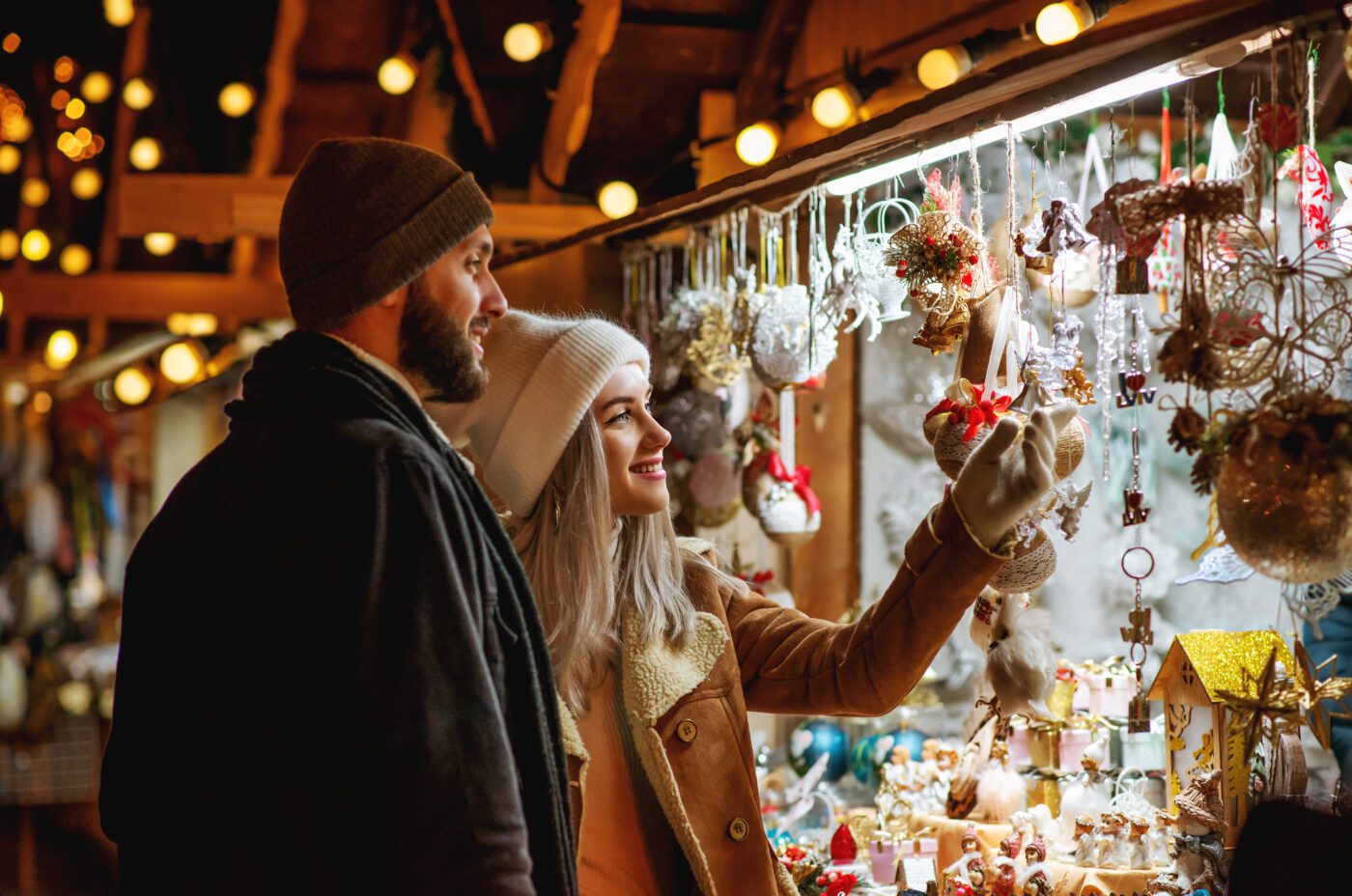 A couple in winter clothes smiles and browses festive ornaments at an outdoor Taste of London holiday market stall lit with warm string lights. The woman points at a decoration while the man stands beside her.