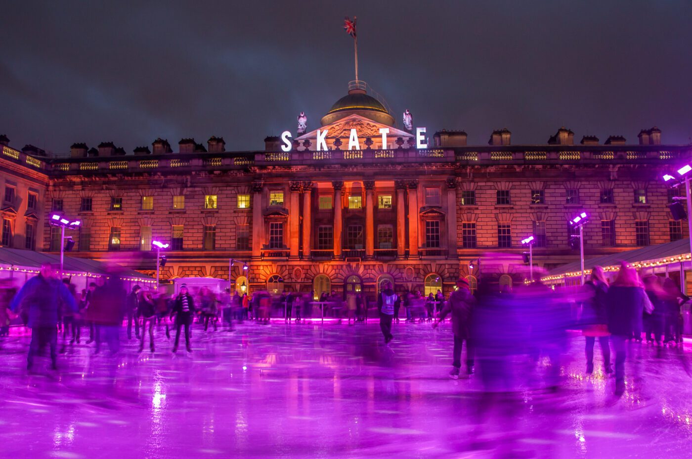 People ice skating at night in front of a grand, illuminated building with a sign that reads 