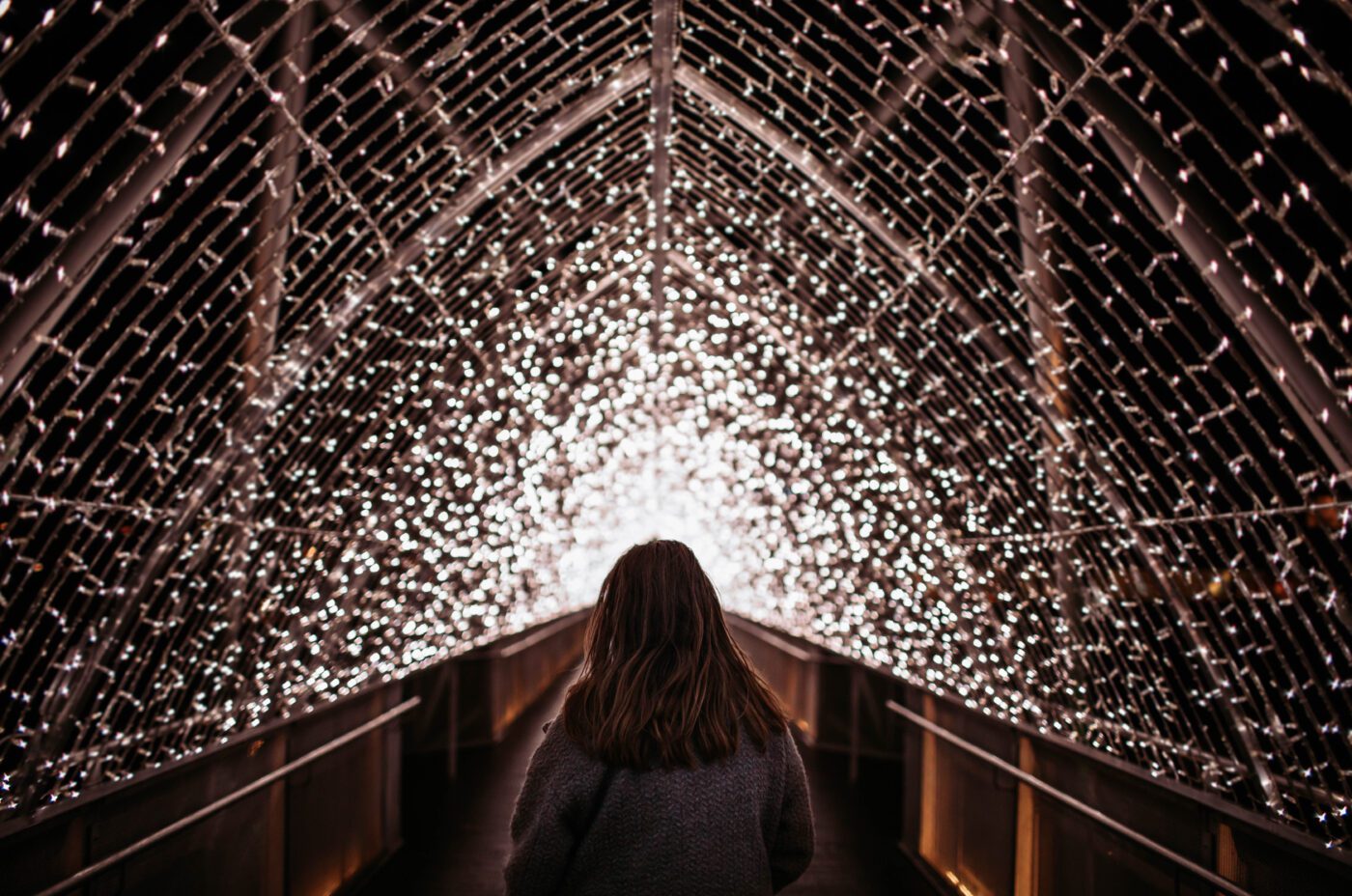 A person with long hair and a coat stands facing a tunnel covered in thousands of small, glowing white lights, creating a bright and dazzling archway effect—perfect for capturing the enchanting spirit of TradFest.