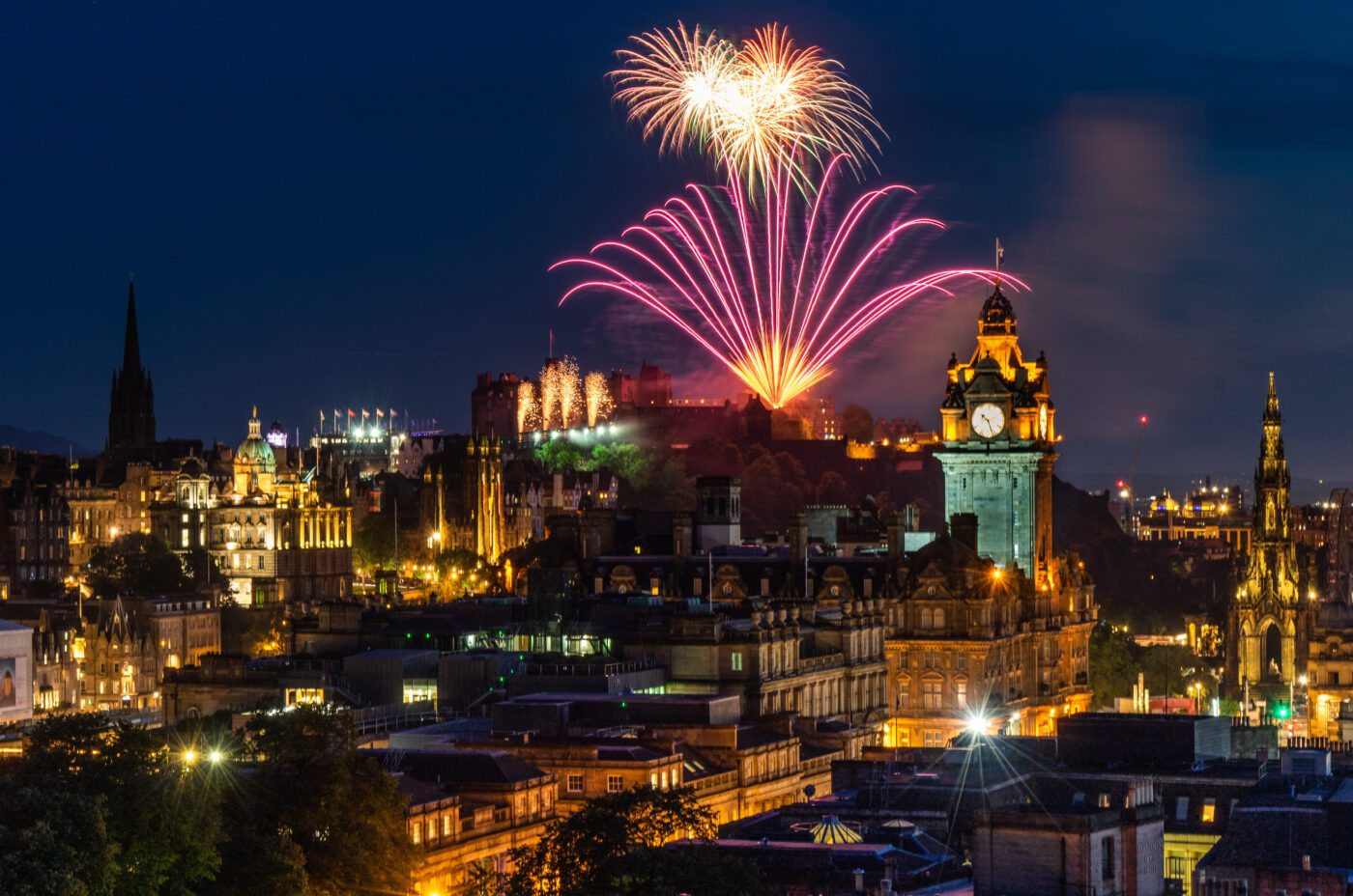 Nighttime cityscape of Edinburgh with bright pink and white fireworks bursting over Edinburgh Castle, historic buildings illuminated, the Balmoral Hotel clock tower in the foreground, and the festive Christmas Market lights sparkling below.