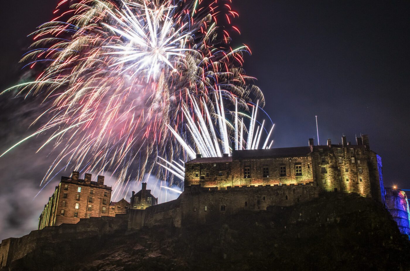 Fireworks light up the night sky above a large, illuminated stone castle perched on a rocky hill, as the nearby Christmas Market adds a festive and dramatic atmosphere.