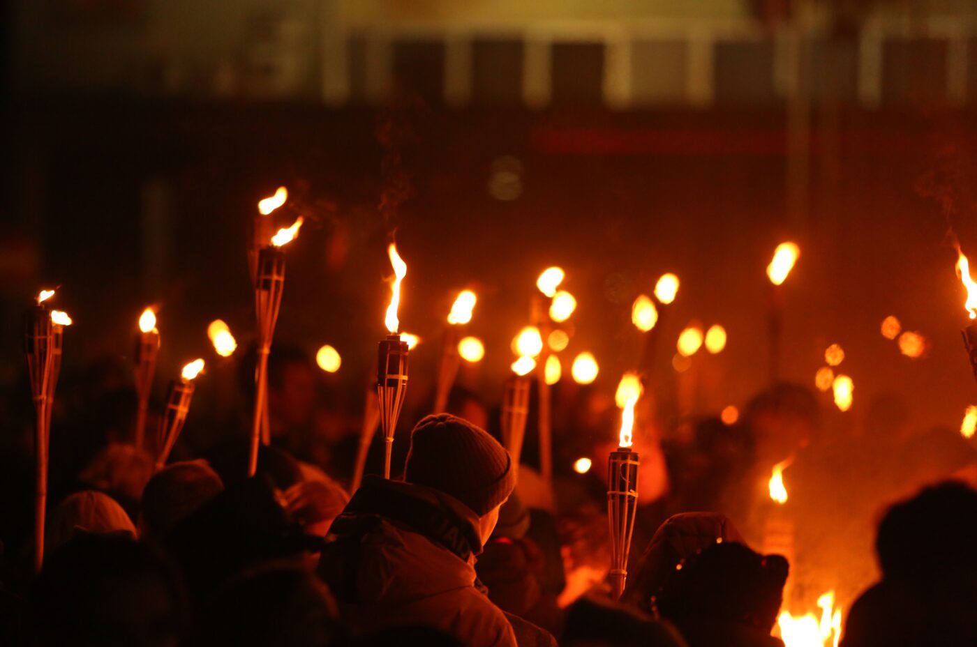 A crowd of people holds lit torches at night, creating a warm glow that illuminates the scene, reminiscent of a festive Christmas Market. The background is dark and the faces in the crowd are mostly obscured.