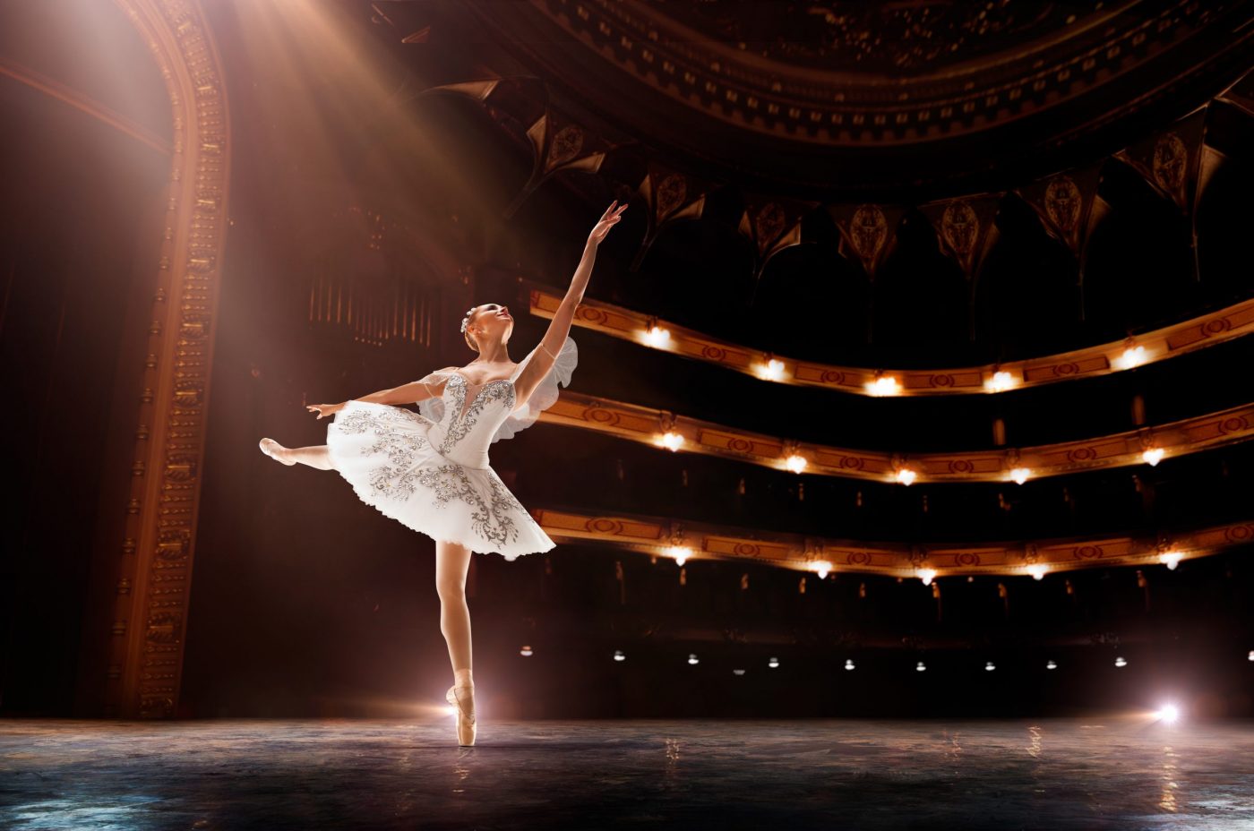A ballerina in a white tutu, radiating pride, strikes an elegant pose onstage under dramatic lighting, with ornate balconies and rows of lights illuminating the grand theater behind her.
