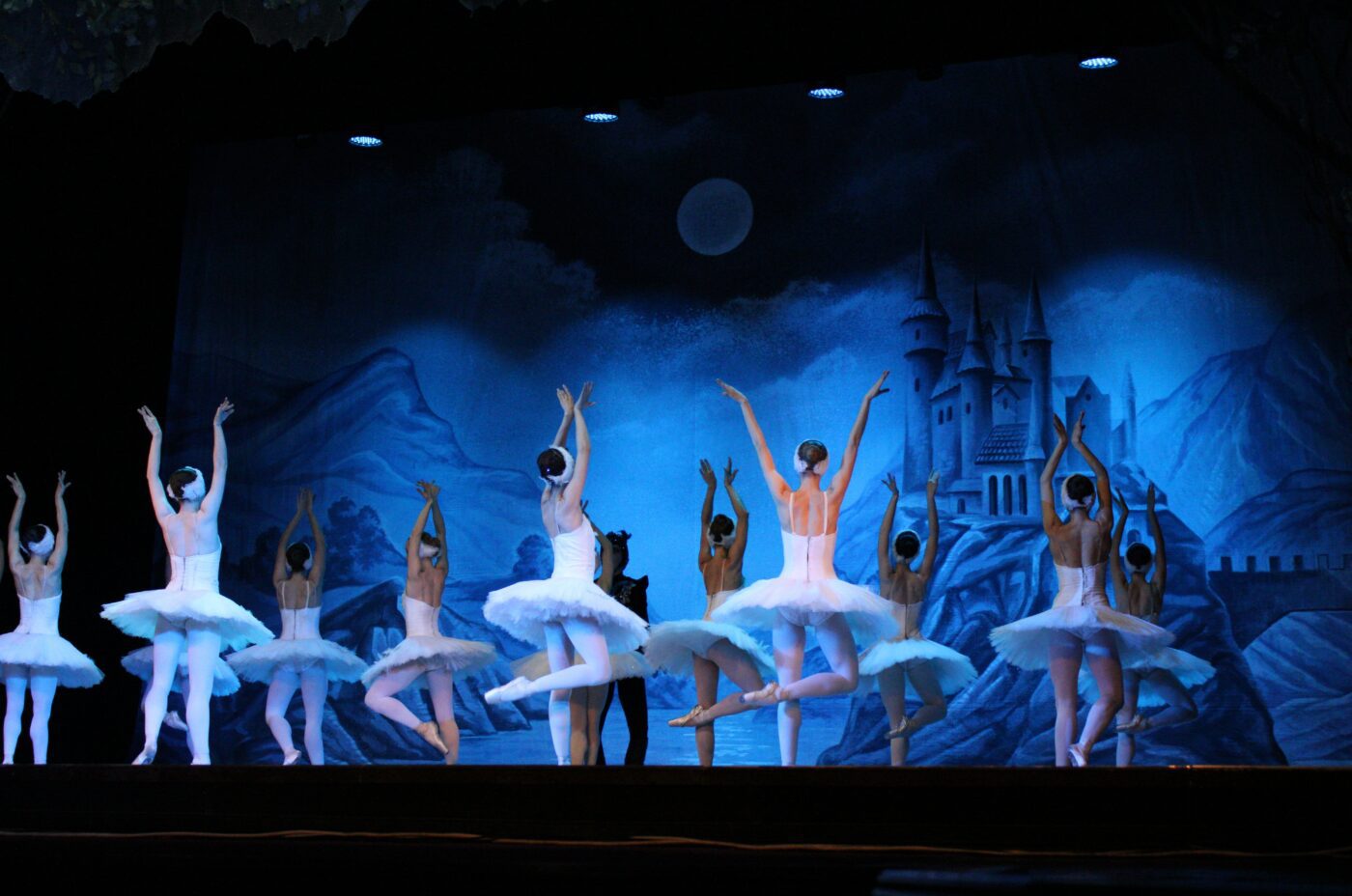 Ballet dancers in white tutus perform on stage with arms raised in pride. A blue-lit backdrop depicts mountains, a full moon, and a castle, creating a dreamy, fairytale atmosphere.