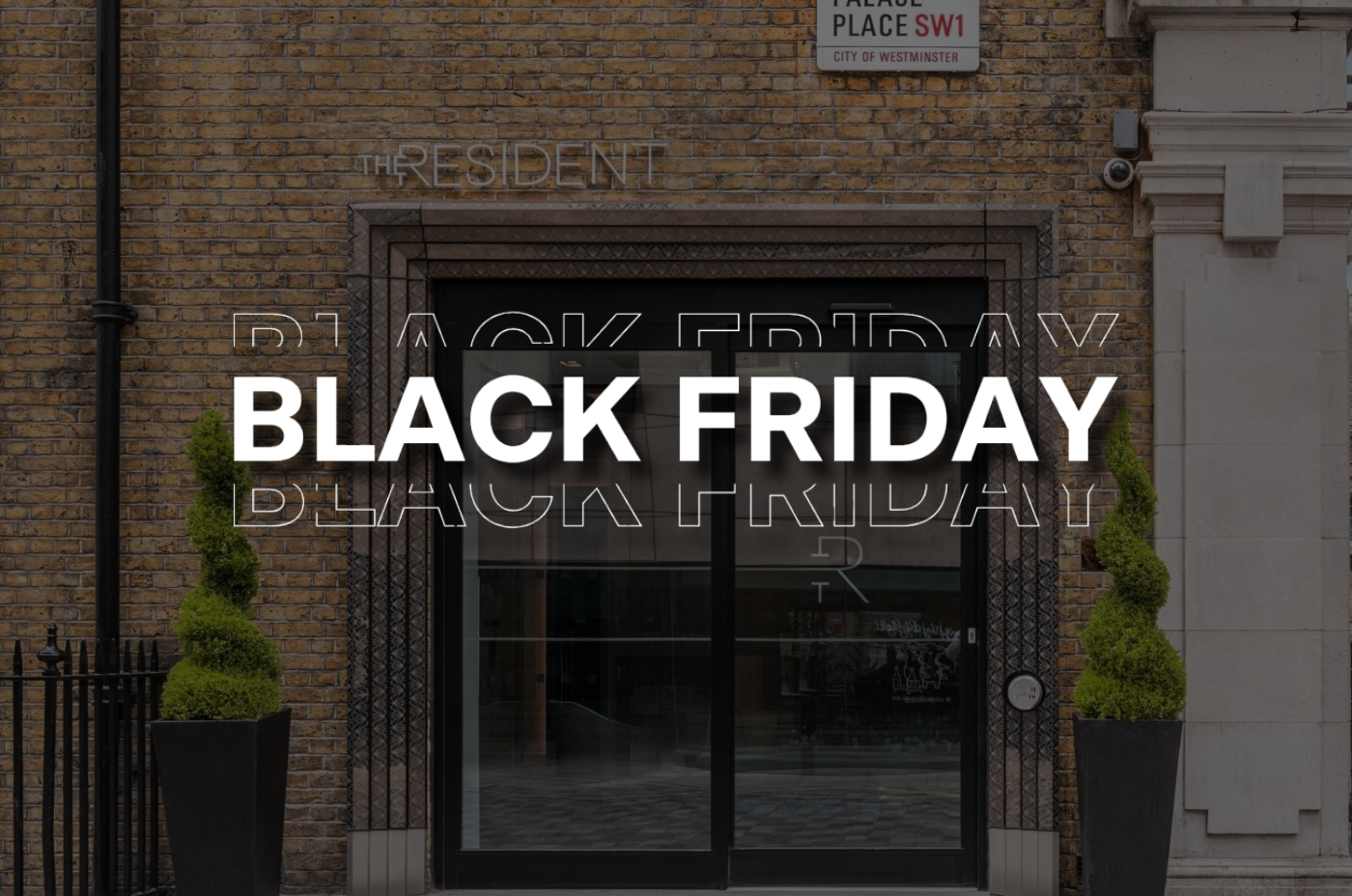 A building entrance with a glass door and two sculpted shrubs, overlaid with large white text reading 