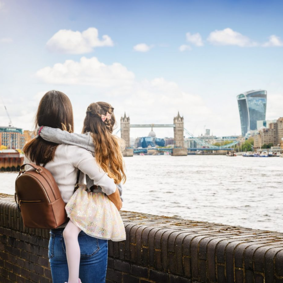 A woman with a brown backpack holds a young girl while looking across the River Thames at Tower Bridge and modern London buildings under a partly cloudy sky.