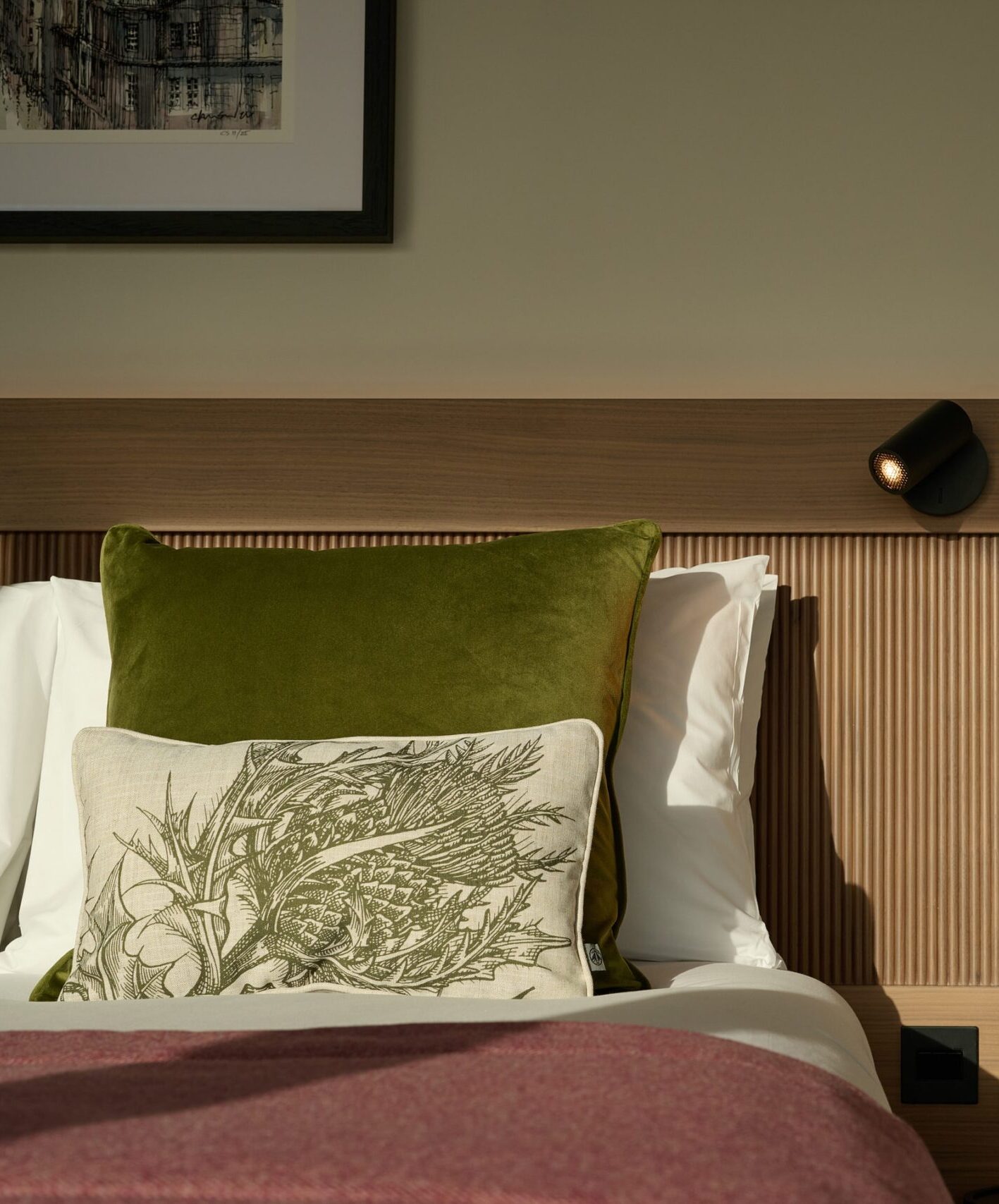 A neatly made bed with white sheets, a green cushion, and a decorative pillow with a botanical design. The wooden headboard features a mounted reading light. Sunlight streams into the modern, cozy room.