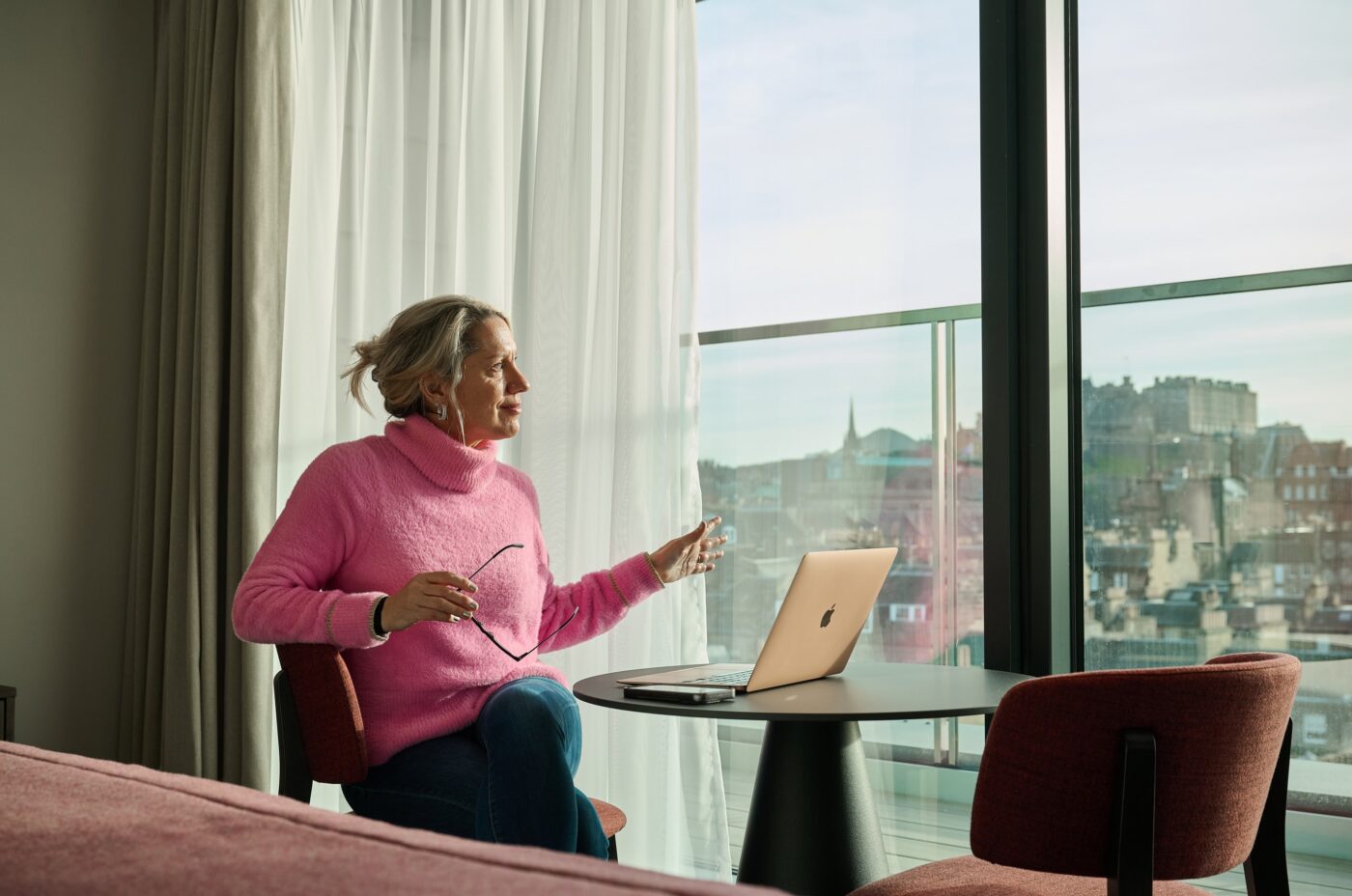 A woman in a pink sweater sits at a table with a laptop, holding glasses, and looks out of a large window at a cityscape. Natural light fills the modern room with floor-to-ceiling curtains.