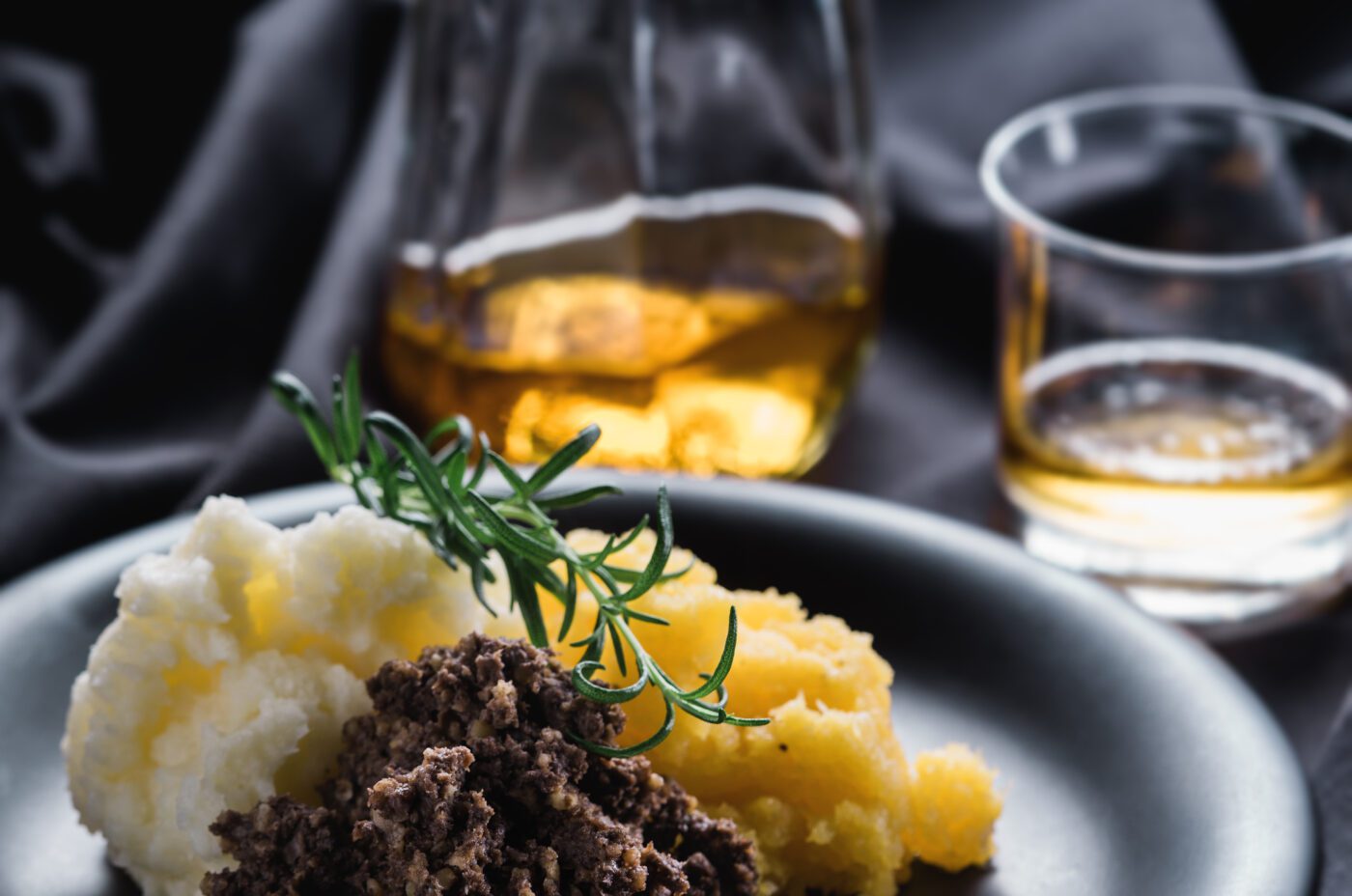 A plate with mashed potatoes, yellow turnips, and a portion of haggis garnished with rosemary evokes the spirit of a highland show, with a glass decanter and whisky in the background on a dark tablecloth.