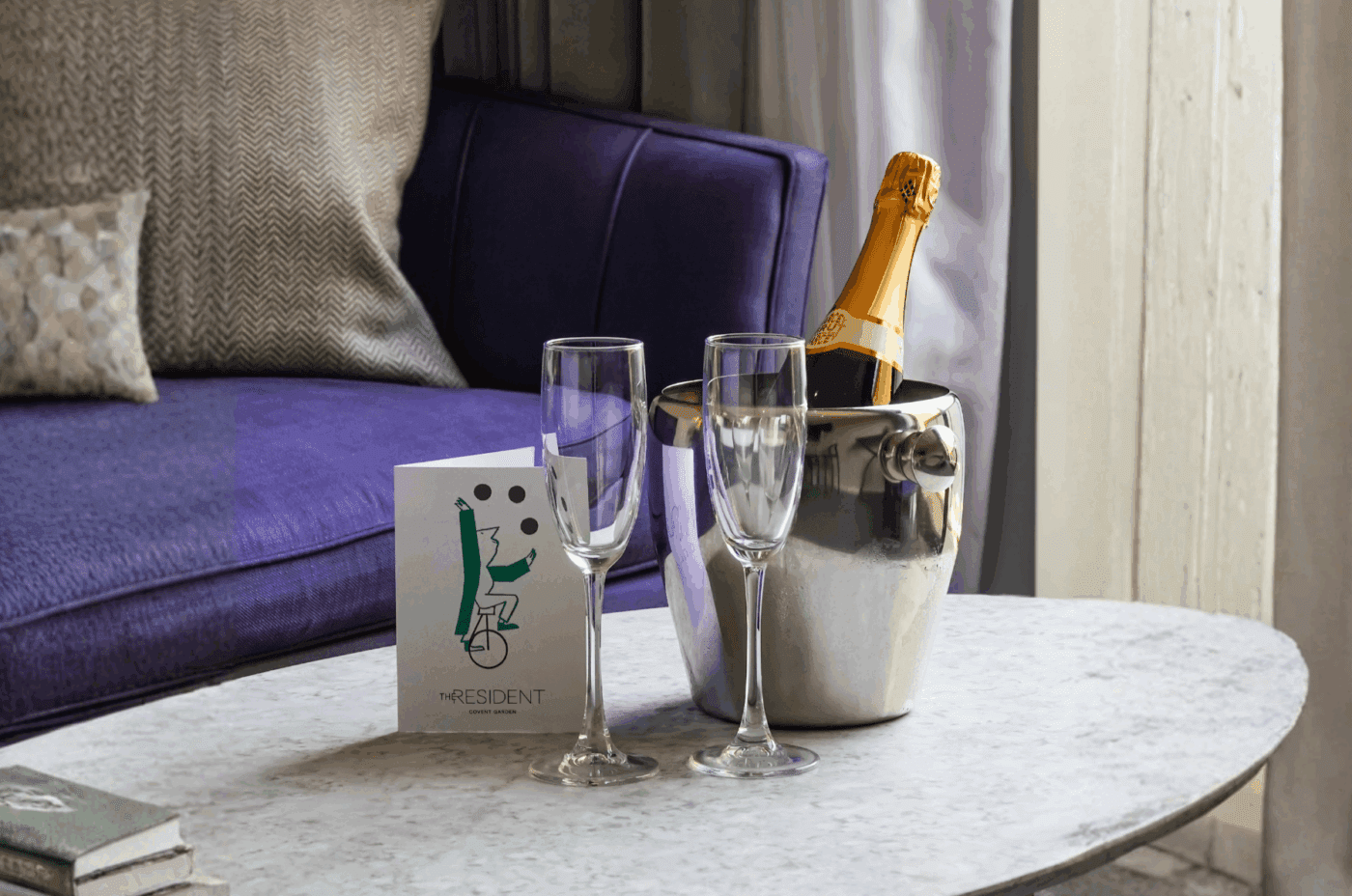A bottle of champagne in an ice bucket, two empty champagne flutes, and a card sit on a marble table in front of a purple sofa with patterned cushions. Sunlight filters through a window with sheer curtains.
