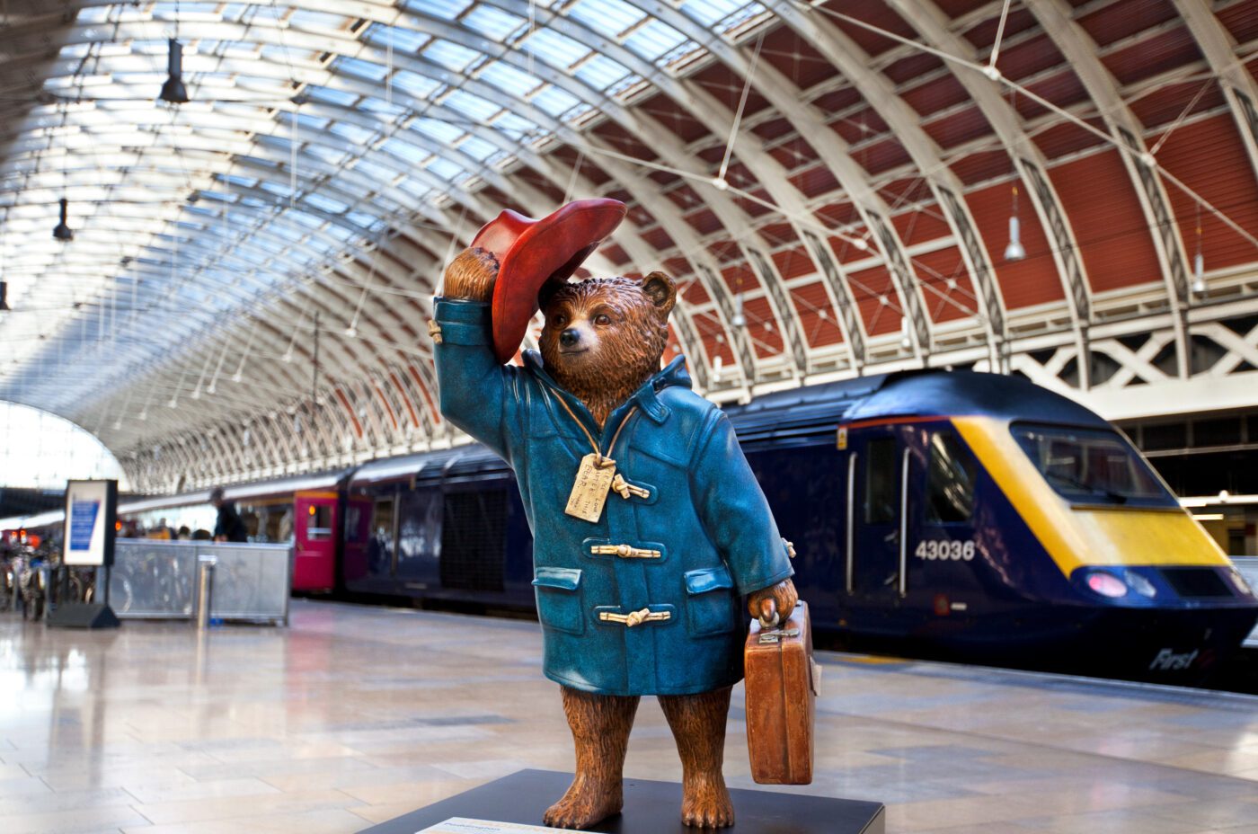 A statue of Paddington Bear in a blue coat and red hat holding a suitcase stands inside a spacious train station, reminiscent of the charm found at Kew Fete, with a modern train visible on the tracks in the background.