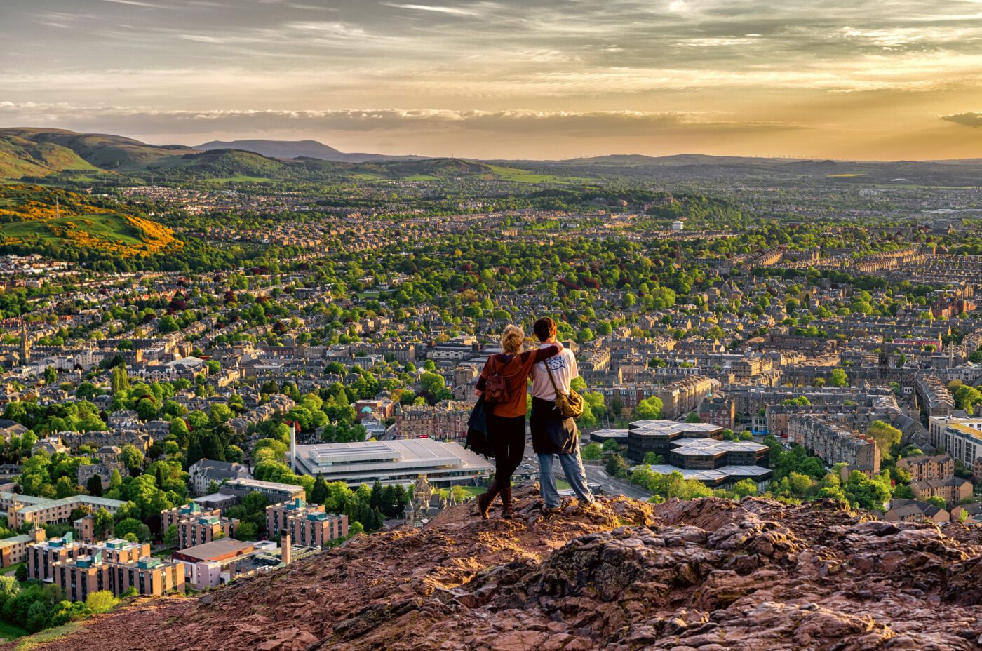 Three people stand on a rocky hilltop at sunset, overlooking the city below, as the International Children's Festival fills the air with energy amid green trees, buildings, and distant hills beneath a partly cloudy sky.